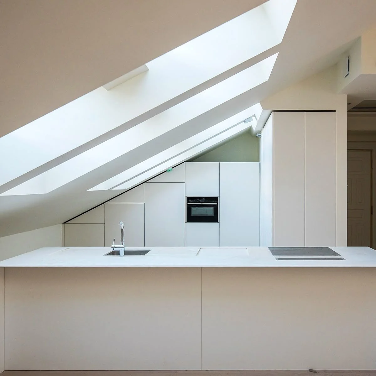 Handleless white kitchen fitted under a sloped ceiling with skylights, island with sink and induction hob